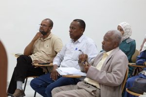 Group of adults seated in a meeting, looking attentive, with papers and pens on shared table.