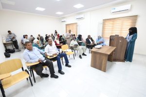 A woman in a hijab stands at a wooden podium presenting to a seated audience in a bright classroom with blinds and ceiling lights.