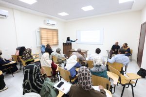 Lecturer at a wooden podium teaching a group of adults seated in a bright classroom with a projected slide on the screen.