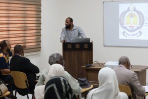 Bearded man in a striped shirt speaks from a wooden podium at the front of a classroom, a projector screen with a logo visible beside him.