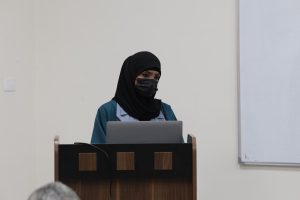 Woman wearing a black hijab and face mask standing at a lectern with a laptop in a classroom setting