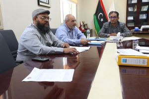 Three men sit at a polished conference table with papers, a water bottle, and flags in the background, in a formal office setting.