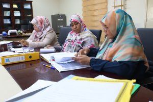 Three women in colorful headscarves sit at a conference table, reviewing documents in a meeting room.