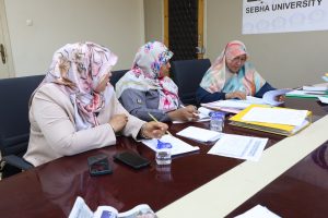 Three women wearing patterned headscarves sit at a conference table reviewing documents and taking notes, with papers and phones in front of them.