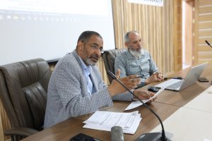 Two men seated at a wooden conference table with microphones, papers, and laptops during a meeting; foreground man speaks while gesturing with hands.