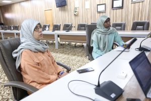 Two women in hijabs sit at a conference table with microphones in a formal meeting room setting.