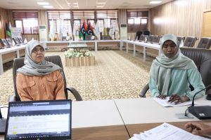 Two women wearing hijabs sit at a large conference table with laptops and papers in a formal meeting room with flags in the background.