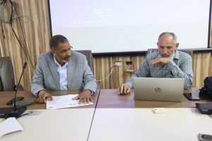 Two men in a meeting sit at a wooden conference table, reviewing documents while one uses a laptop and the other gestures over papers in front of a projector screen.