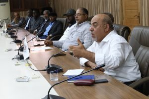 Group of men sitting along a conference table with microphones during a meeting, viewed in an office setting.