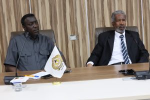 Two men sit at a conference table with microphones, a flag, and papers in front of them, wood panel backdrop behind.
