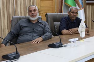 Two men sit at a wooden conference table with microphones, in a formal meeting room with flags in the background.