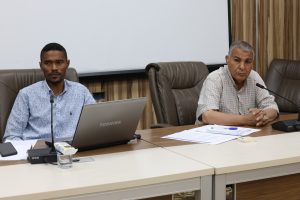 Two men sit at a conference table with a laptop, documents, and microphones in front of them, appearing to participate in a meeting.