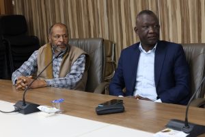 Two men seated at a conference table with microphones; one in a plaid shirt and scarf, the other in a blue suit, against a wood-paneled wall.