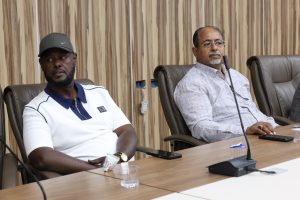 Two men seated at a conference table with microphones, wood-paneled wall behind them.