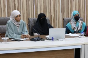 Three women wearing hijabs sit at a conference table with a laptop, microphone, and papers in front of them.
