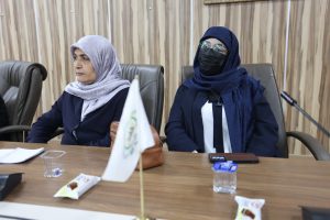 Two women wearing headscarves sit at a conference table in a meeting room, attentive and focused on the discussion.