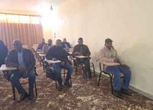 Adult men sit in a classroom at individual desks in rows, some with notebooks, beige walls and a curtain on the left.