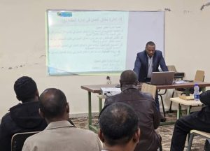 Male presenter in a suit stands at a desk with a laptop, giving a lecture to a classroom; a projected slide with Arabic text is on the wall behind him.