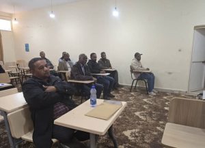 Adult students sit in a small classroom with attached desks, listening to a speaker at the front.