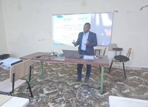 Man in a suit giving a lecture in a sparse classroom, standing behind a desk with a laptop and papers while a projector screen shows slides behind him.