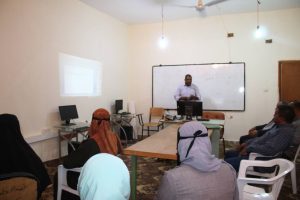Male instructor stands at a lectern presenting to adults in a classroom, with a whiteboard and projection screen behind him.