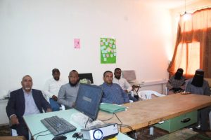 Group of men seated around a long table in a small office, with a computer and projector on the desk and color posters on the wall