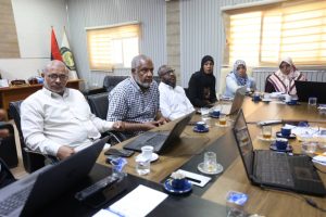 Group of six professionals seated around a wooden conference table with laptops and cups in a meeting room, flags in the background.