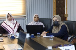 Three women sit around a conference table in a meeting room, one wearing a hijab and a face mask, others looking at a laptop and notes while a notebook and cups are on the table.