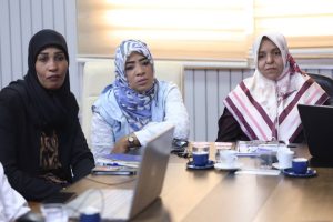 Three women in headscarves sit at a conference table with laptops and coffee cups, looking attentive during a meeting.