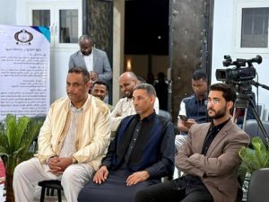 b4060c97-9104-4d04-96a4-b4d827648c41 - جامعة سبها | Sebha University Group of men seated at a formal indoor event; a video camera on a tripod looms to the right while a banner stands in the background.