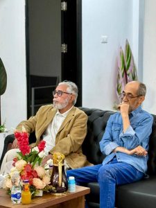 c9bfcc26-76c4-479c-b911-9f17f209ceb2 - جامعة سبها | Sebha University Two older men sit on a black leather sofa in a bright room, one in a beige blazer and white shirt, the other in a denim shirt, with a flower arrangement and drinks on the table nearby.