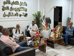 f752bd25-4697-4401-b436-5e8d4c9b95d7 - جامعة سبها | Sebha University Group of men seated in a formal meeting room with an Arabic inscription on the wall, plants, and flags in the background.