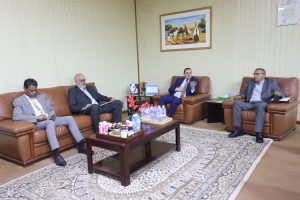 01F891AC-9744-45D6-B4CB-1E1D4441344F - جامعة سبها | Sebha University Four men in suits sit on brown leather couches in a meeting room; table holds drinks and snacks, with small flags and a camel painting on the wall behind them.