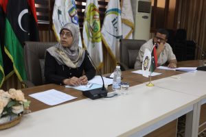 Two people sit at a meeting table with documents, microphones, and flags in the background.