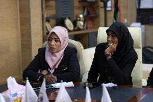 Two women wearing hijabs sit at a conference table; one in pink writes while the other in black looks on, in a meeting room.