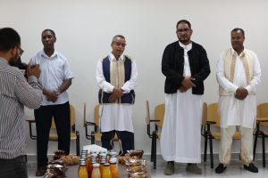 Photographer taking a photo of five men in traditional clothing standing behind a row of chairs, with snacks and drinks on the table in front.
