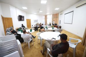 0F7A29E2-DCB8-426F-9DA1-91681712E290 - جامعة سبها | Sebha University A conference room with people seated around round tables, facing a panel at the front and a projector screen on the right wall.