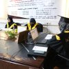 Three women in black graduation gowns and yellow sashes sit at a conference table with laptops and notebooks, a banner in Arabic hangs behind them.