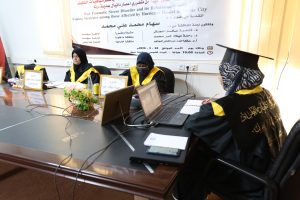 Three women in black graduation gowns and yellow sashes sit at a conference table with laptops and notebooks, a banner in Arabic hangs behind them.