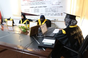 Three women in black graduation gowns and yellow sashes sit at a conference table with laptops and notebooks, a banner in Arabic hangs behind them.