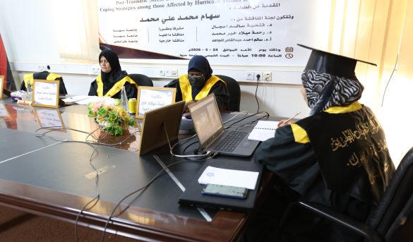 177D0280-E121-42CA-A0E0-4E4098A3AD8C - جامعة سبها | Sebha University Three women in black graduation gowns and yellow sashes sit at a conference table with laptops and notebooks, a banner in Arabic hangs behind them.