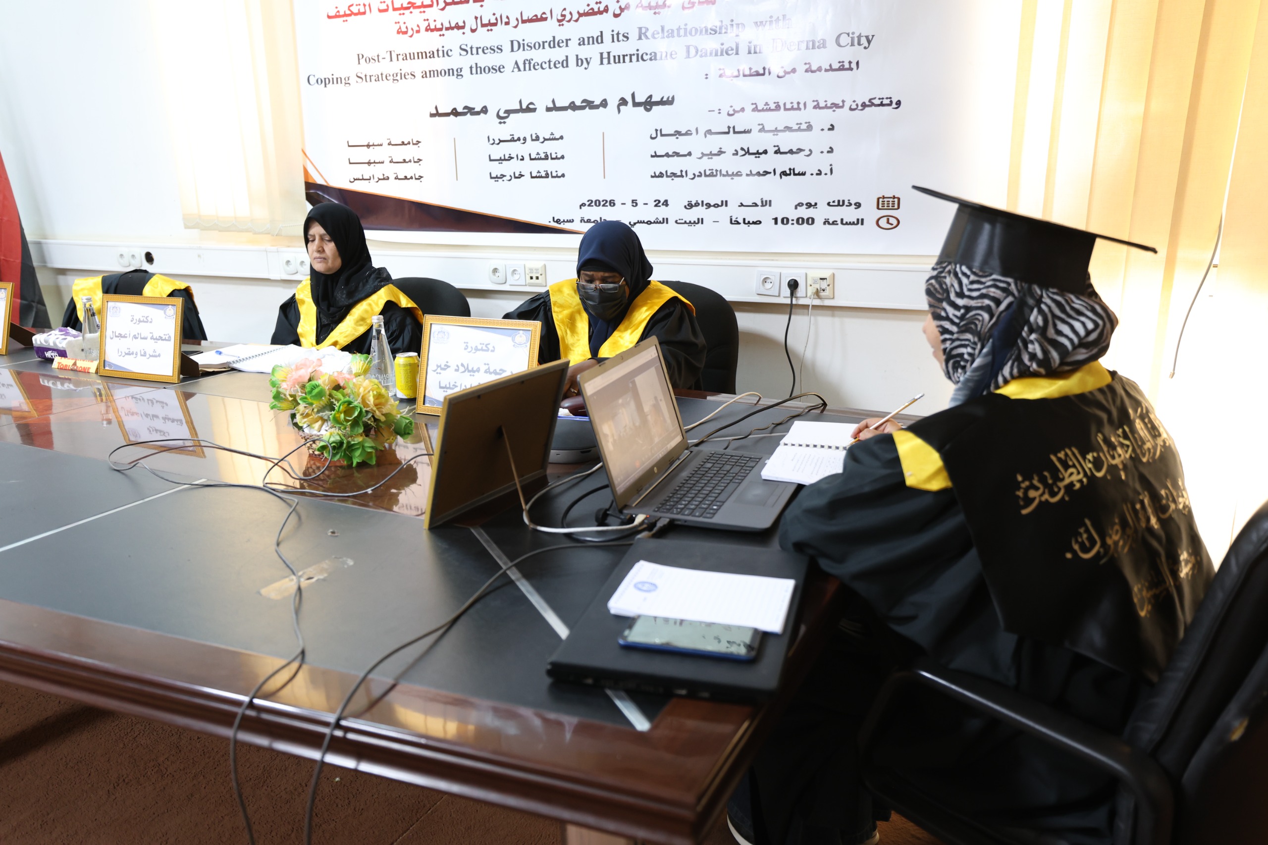 Three women in black graduation gowns and yellow sashes sit at a conference table with laptops and notebooks, a banner in Arabic hangs behind them.