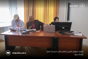 1952C92B-D393-44B7-BE7A-33DBA63CFA66 - جامعة سبها | Sebha University Three people sit at a conference table with laptops and papers; a man in a cap on the left, a center man in a suit, and a woman in a hijab on the right, in a room with yellow curtains and a university logo on the wall.