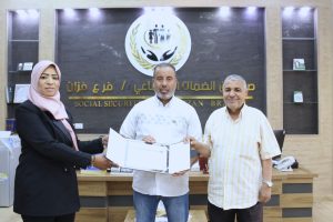 Three people stand in an office holding a certificate, with a social-security emblem and Arabic signage behind them.