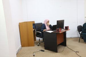 1D1913F0-3827-411E-A185-D82DAD67D13B - جامعة سبها | Sebha University Woman in a pink hijab sits at a desk with a laptop, monitor, and red speakers in a cluttered office space.