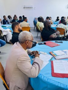1F7E7F67-3773-4DA0-9FC1-99378F6555EB - جامعة سبها | Sebha University A conference room with attendees seated at blue-tablecloth tables; foreground man with gray hair writes in a notebook.