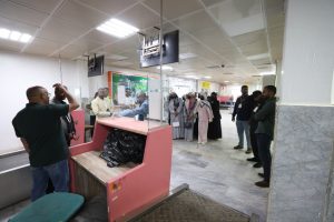 People in line at a security checkpoint inside a building; a pink screening table and overhead monitors are visible ahead of them.