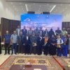 Group of men and women posing for a group photo on a conference stage with a blue banner behind them.