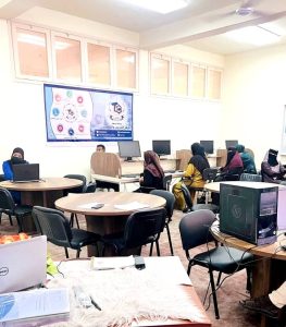 Classroom computer lab with students seated at round tables using laptops, many wearing hijabs.