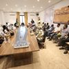 Group of men and women seated around a long conference table in a meeting room, with a Libyan flag and a banner on the wall behind them.